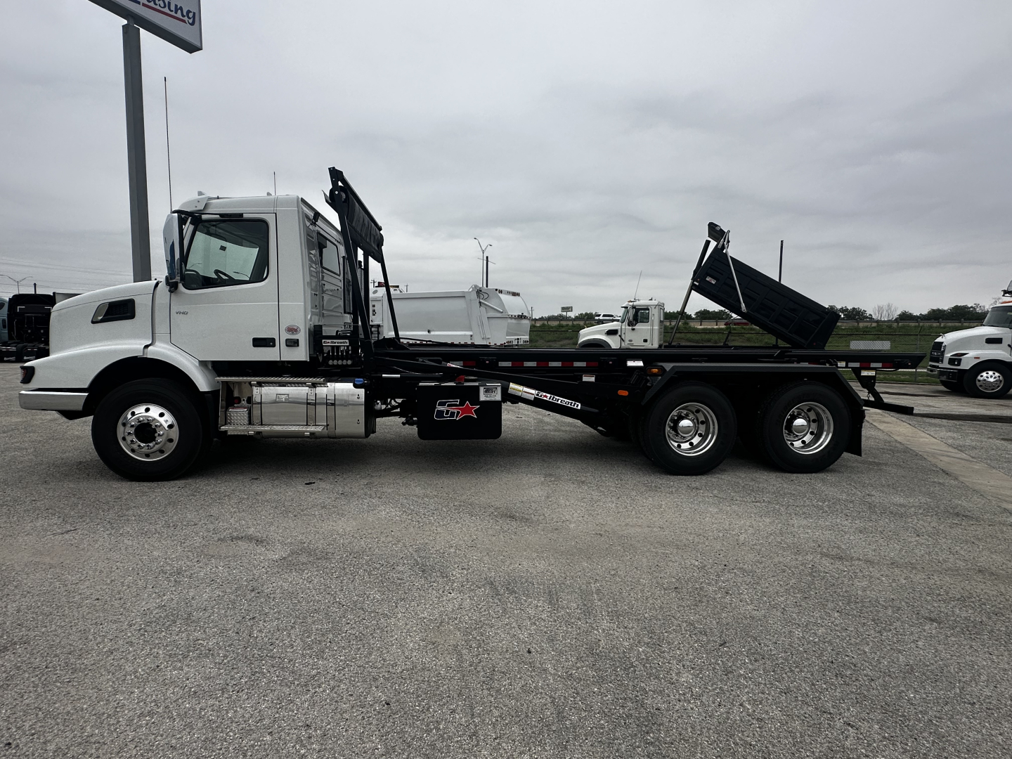 Volvo VHD64B300 roll-off truck side view showing flatbed, hydraulic hoists, and dual rear axles.