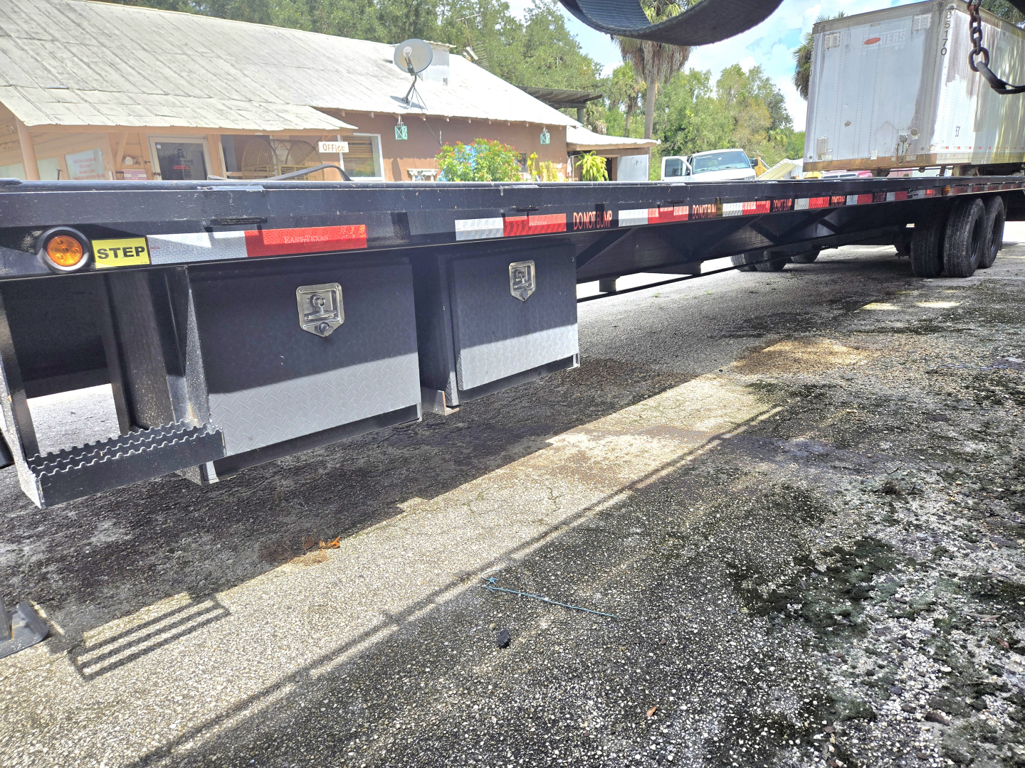 Flatbed trailer undercarriage showing toolbox boxes, steps, and rear axles with reflective tape. Side rail shows caution stickers and decals; truck yard in background.