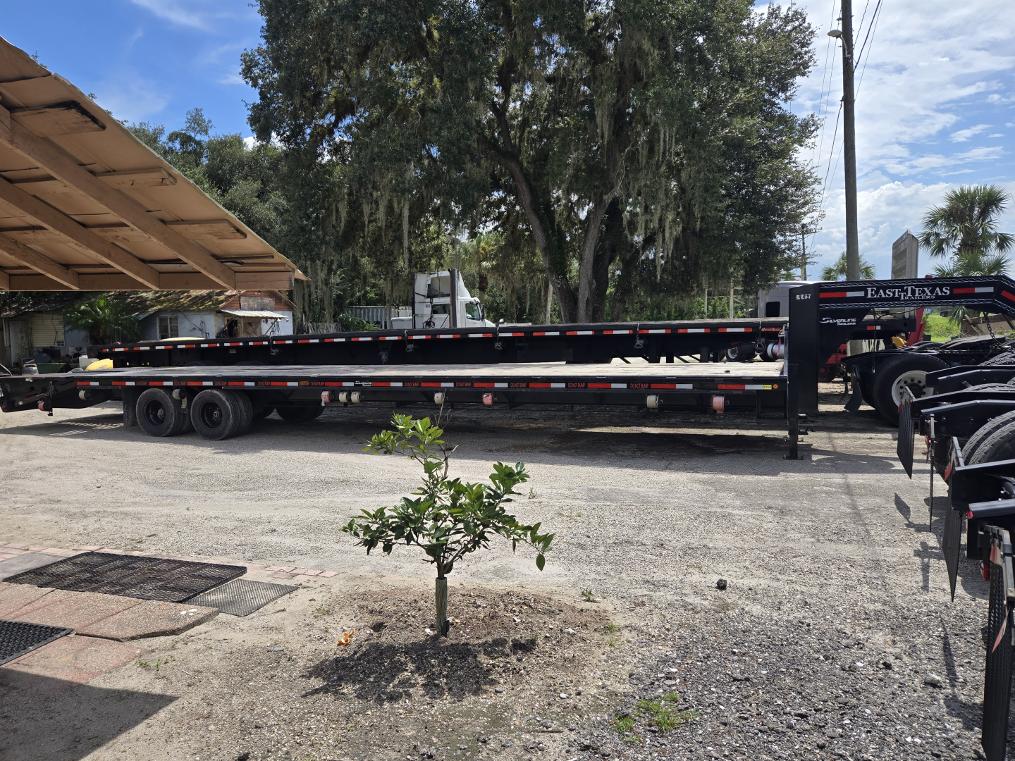 Flatbed trailer with gooseneck and ramps, long black trailer with dual axles and striped safety markings, rear ramps visible under a shaded area.