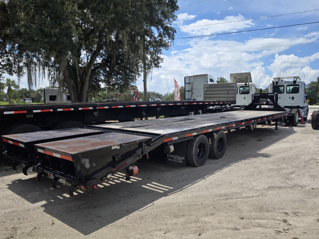 Flatbed trailer with long deck and ramps, three-axle with black frame and red-and-white safety markings, parked in lot with trucks.