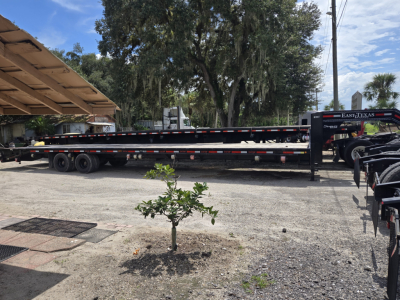Flatbed trailer with gooseneck and ramps, long black trailer with dual axles and striped safety markings, rear ramps visible under a shaded area.