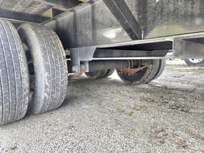 Trailer undercarriage showing dual rear axles and undercarriage components with tires and gravel surface.