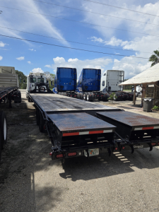 Flatbed trailer with gooseneck 40 ft trailer and ramps, multiple blue trucks in background, sunny day, gravel lot.