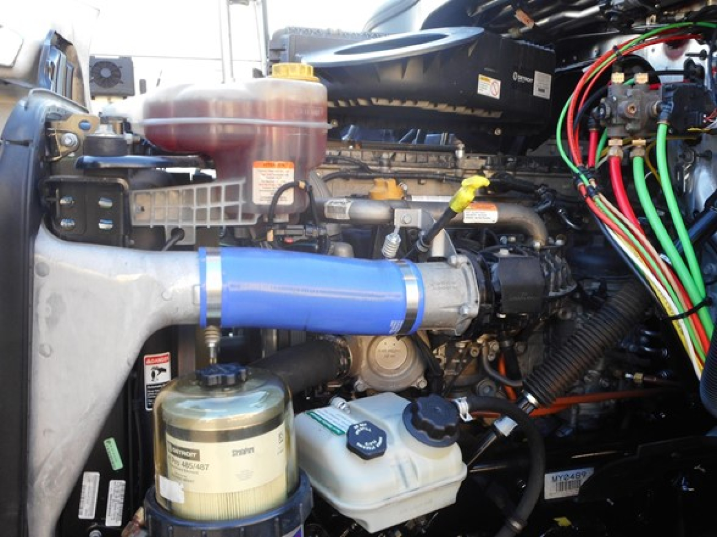 Truck engine bay with blue intercooler pipe, coolant reservoir, power steering/engine fluids visible, assorted hoses and wiring in Freightliner Cascadia engine bay