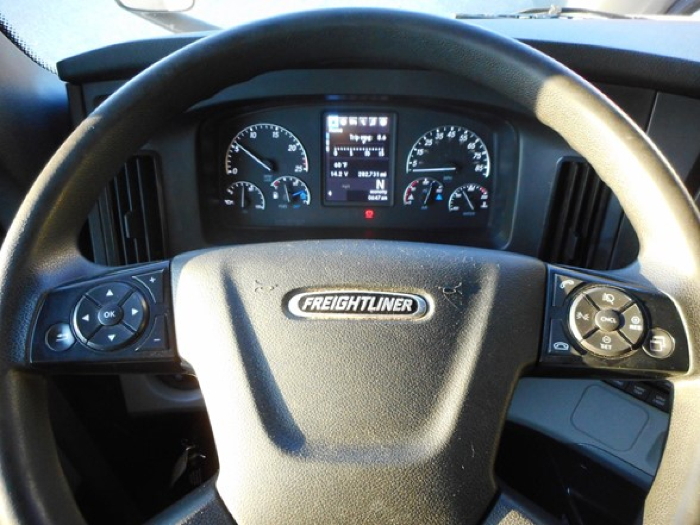 Steering wheel view of a Freightliner truck cockpit with gauges and digital display behind steering wheel, Freightliner emblem visible on center.