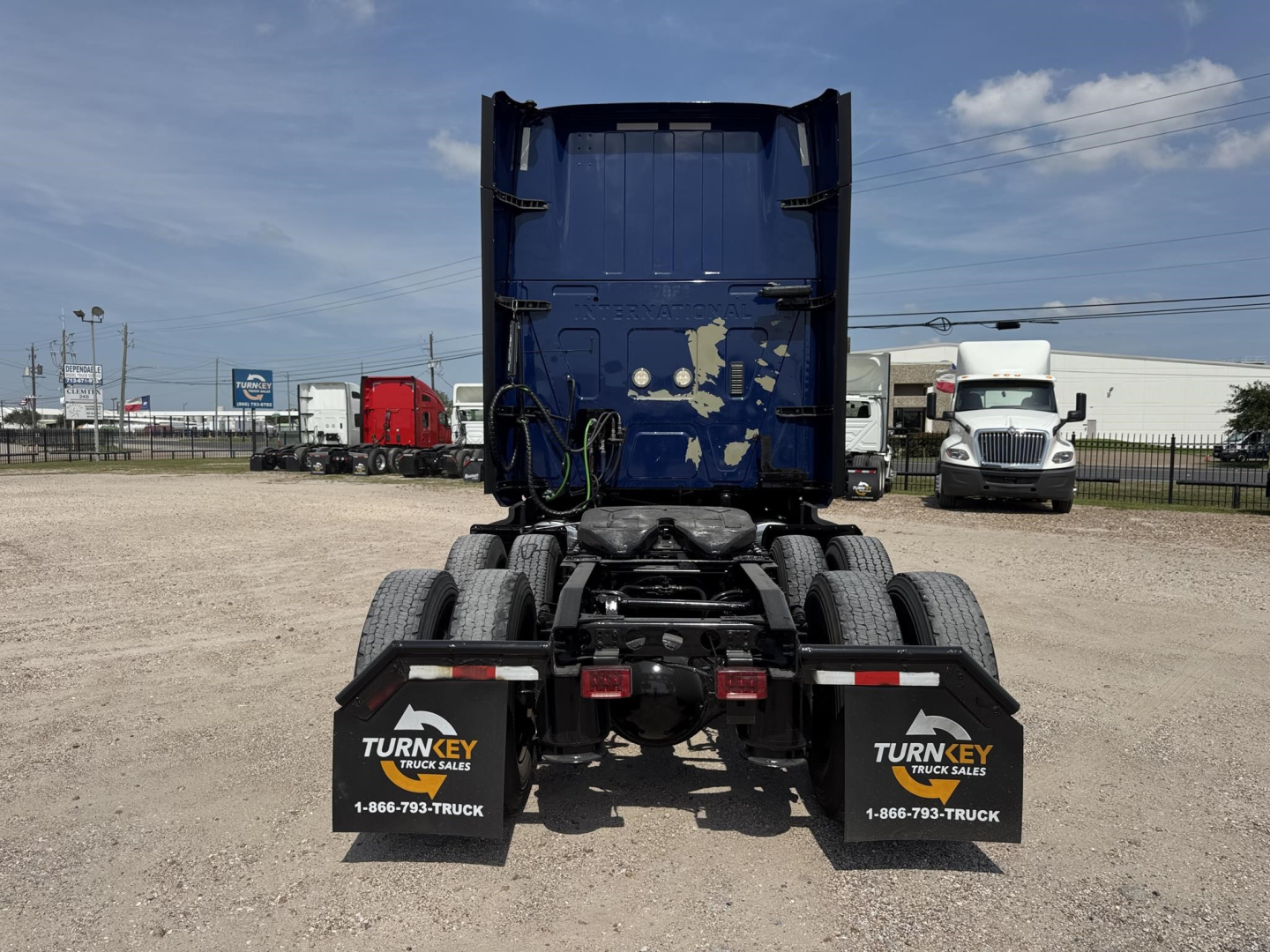 Truck chassis rear view showing fifth wheel kingpin, dual rear axle with mud flaps, and blue sleeper cab in the background; TurnKey Truck Sales branding on mud flaps.