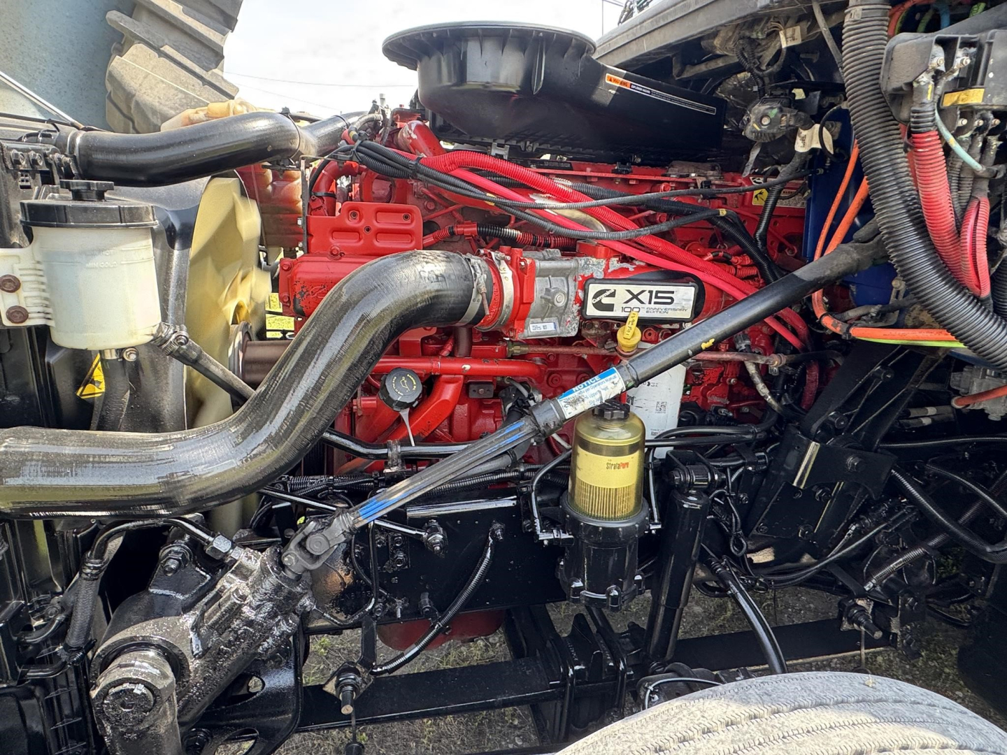 Truck engine bay with bright red Cummins X15 engine block, black hoses, yellow oil filter, and surrounding black chassis components.