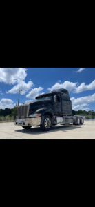 Peterbilt 387 truck shown in side/front view. Black cab with sleeper, chrome bumper, dual rear axles, visible fuel tanks and steps. Clear sky and concrete lot.