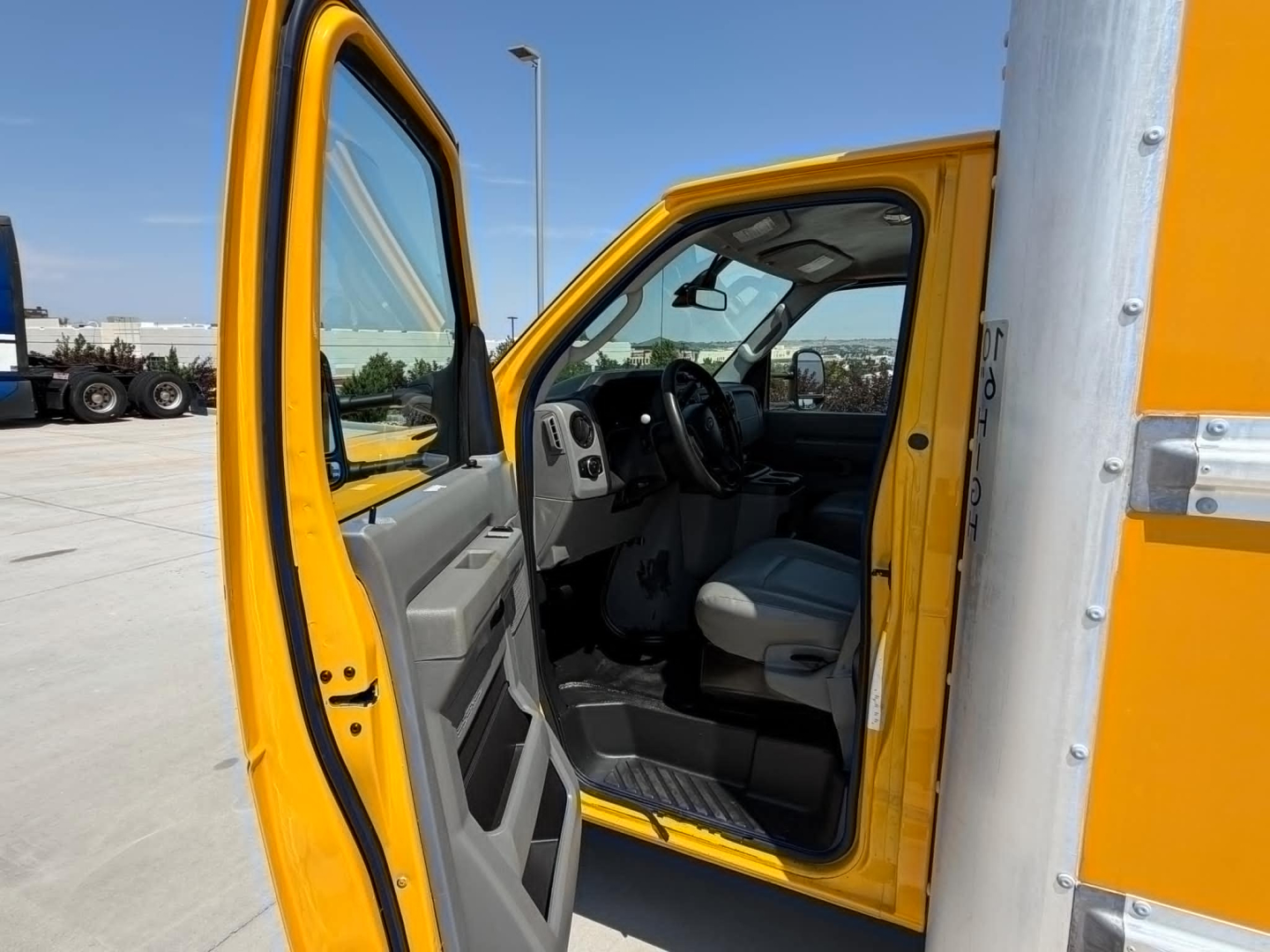 Ford E-Series 2022 cutaway cargo van interior view with yellow exterior, driver door open showing steering wheel, dashboard, and gray seating.