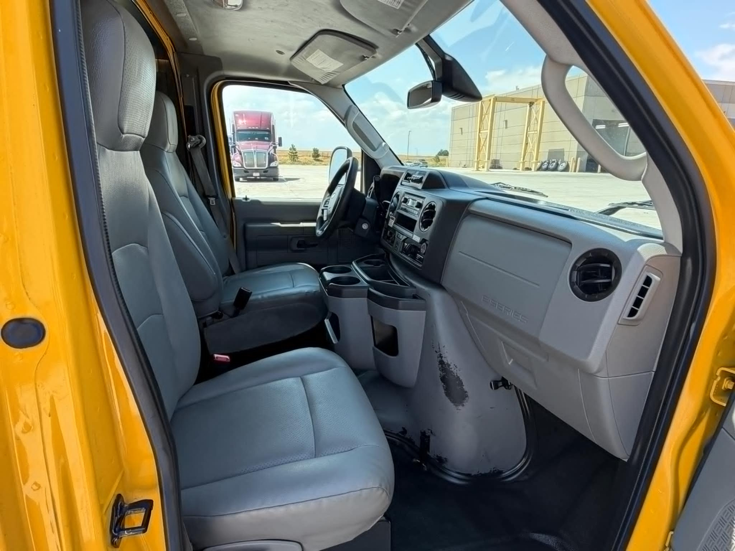 Ford E-Series van interior showing front seats and dashboard; grey cloth seats, steering wheel, central console, and driver-side door; yellow exterior visible on the door frame.