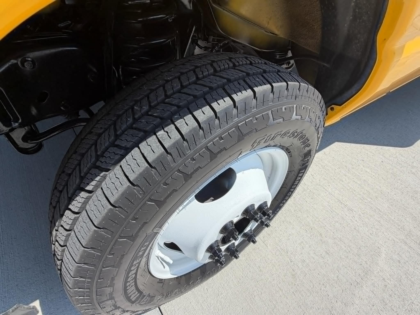 Ford E-Series wheel and fender area showing a tire with alloy wheel and hub, part of a yellow cargo van. Visible tread pattern and wheel nuts.