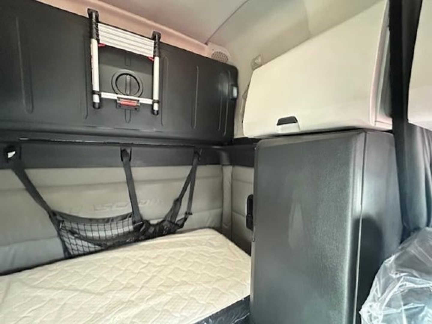 Truck sleeper cab interior with storage rack and netting, quilted mattress visible, overhead compartments, light gray and black trim.