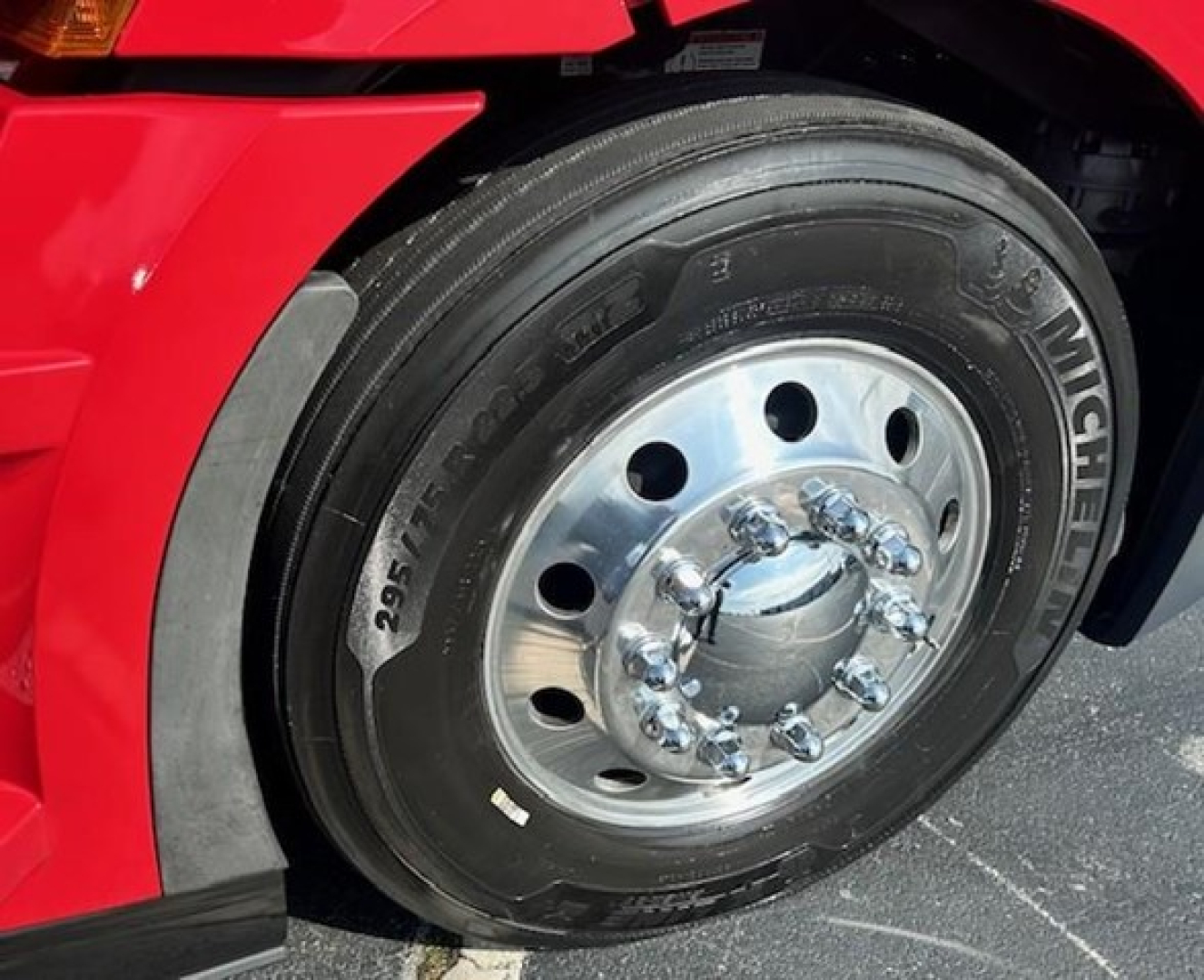 Wheel area of a red Freightliner Cascadia showing Michelin tire and chrome hubcap, with lug nuts visible.