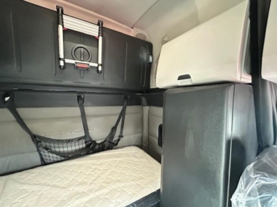 Truck sleeper cab interior with storage rack and netting, quilted mattress visible, overhead compartments, light gray and black trim.
