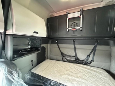 Interior sleeper cab of a Freightliner Cascadia 126 showing a bed with quilted cover, overhead storage cabinet, and mounted equipment.