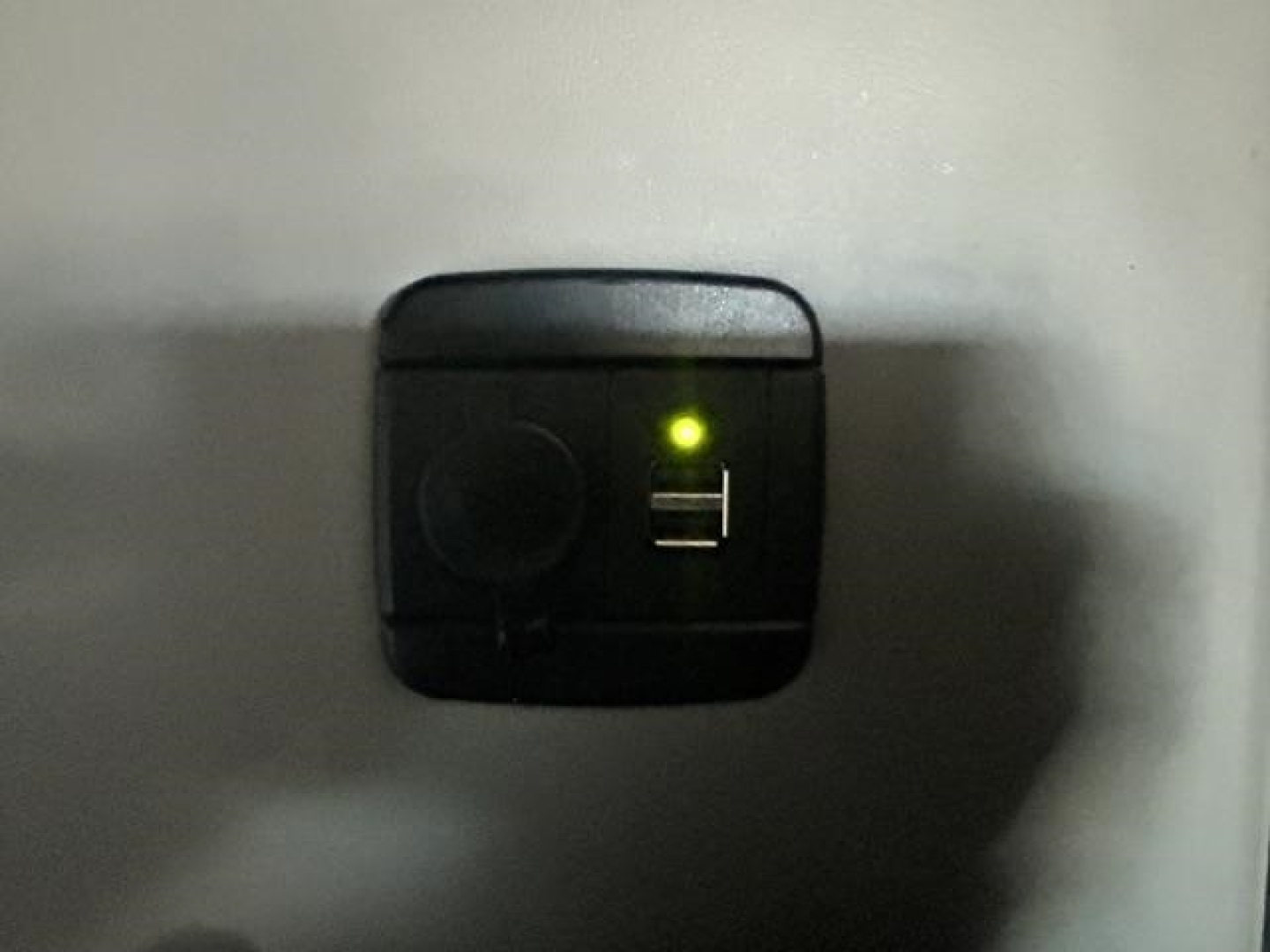 Small black 12V auxiliary power port with a green LED indicator and a USB port visible on a metal surface.
