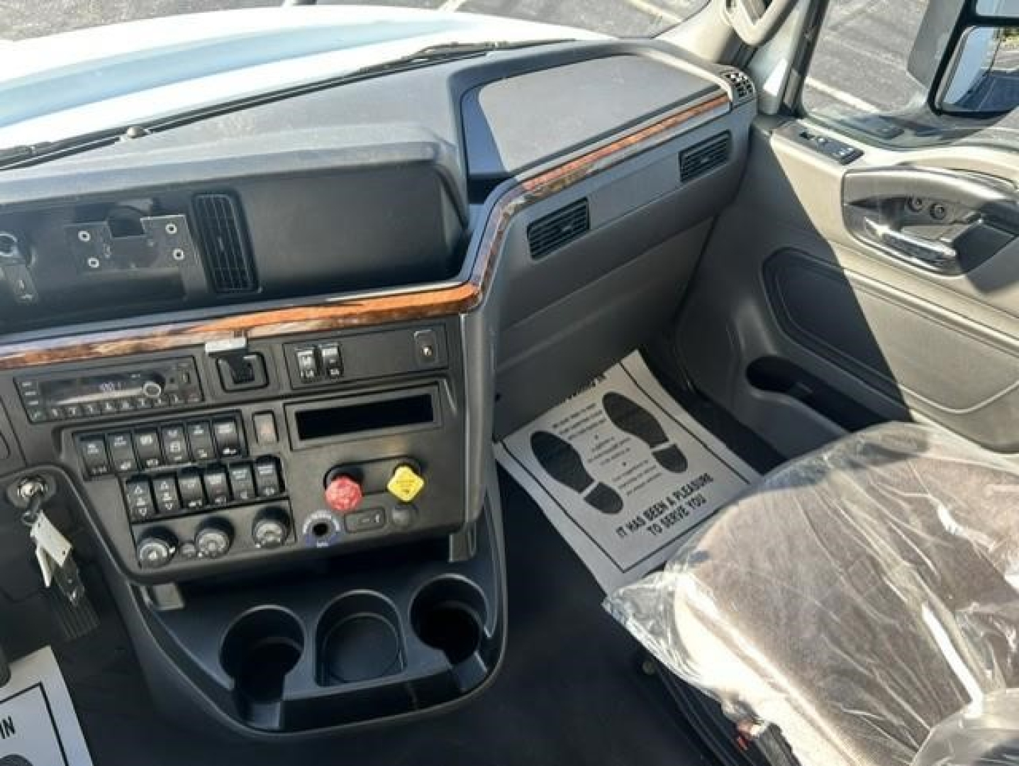 Truck cab interior showing dashboard with switches, wood trim, steering column, and 3 cup holders; floor mat with text on passenger side.