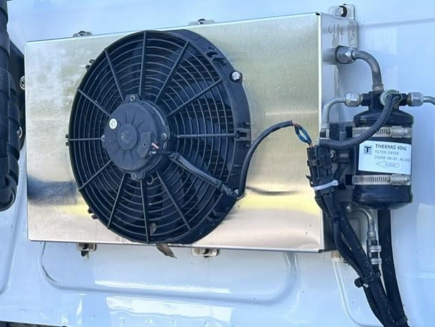 Image shows a cooling module with a large round radiator fan and a Thermo King component mounted on a metallic panel, on the side of a white truck cab.