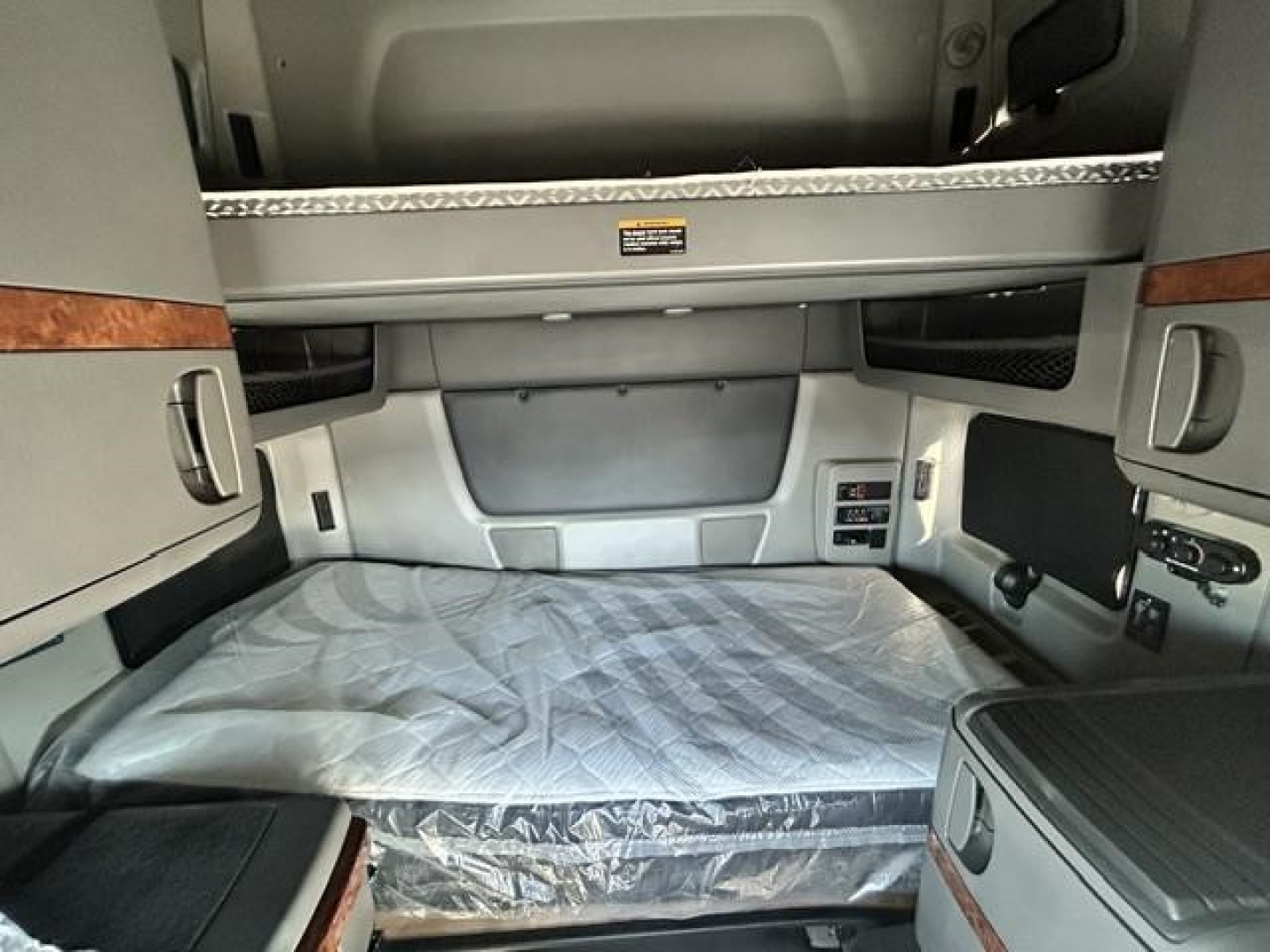 Interior of a truck sleeper cab with a bed mattress in plastic cover, overhead storage cabinets, and control panels on the side.