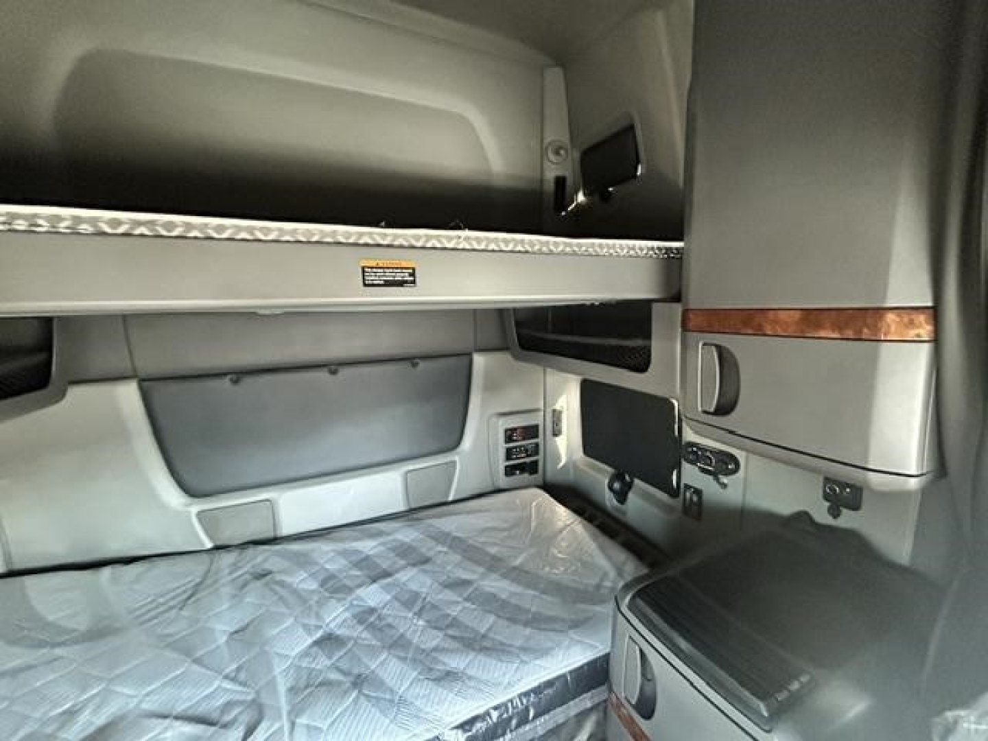 Cab interior of a truck sleeper showing bunk area and side panel with storage, grey upholstery, and metal edge trim.