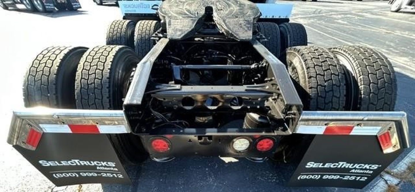 Rear view of a truck chassis showing dual rear axles with heavy-duty tires and a hitch assembly. Visible text on mudflaps reads 'SELECTTRUCKS' and a phone number. No other legible labels.