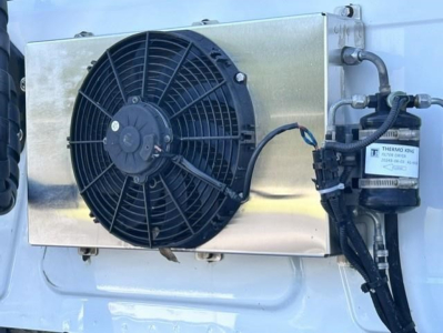 Image shows a cooling module with a large round radiator fan and a Thermo King component mounted on a metallic panel, on the side of a white truck cab.