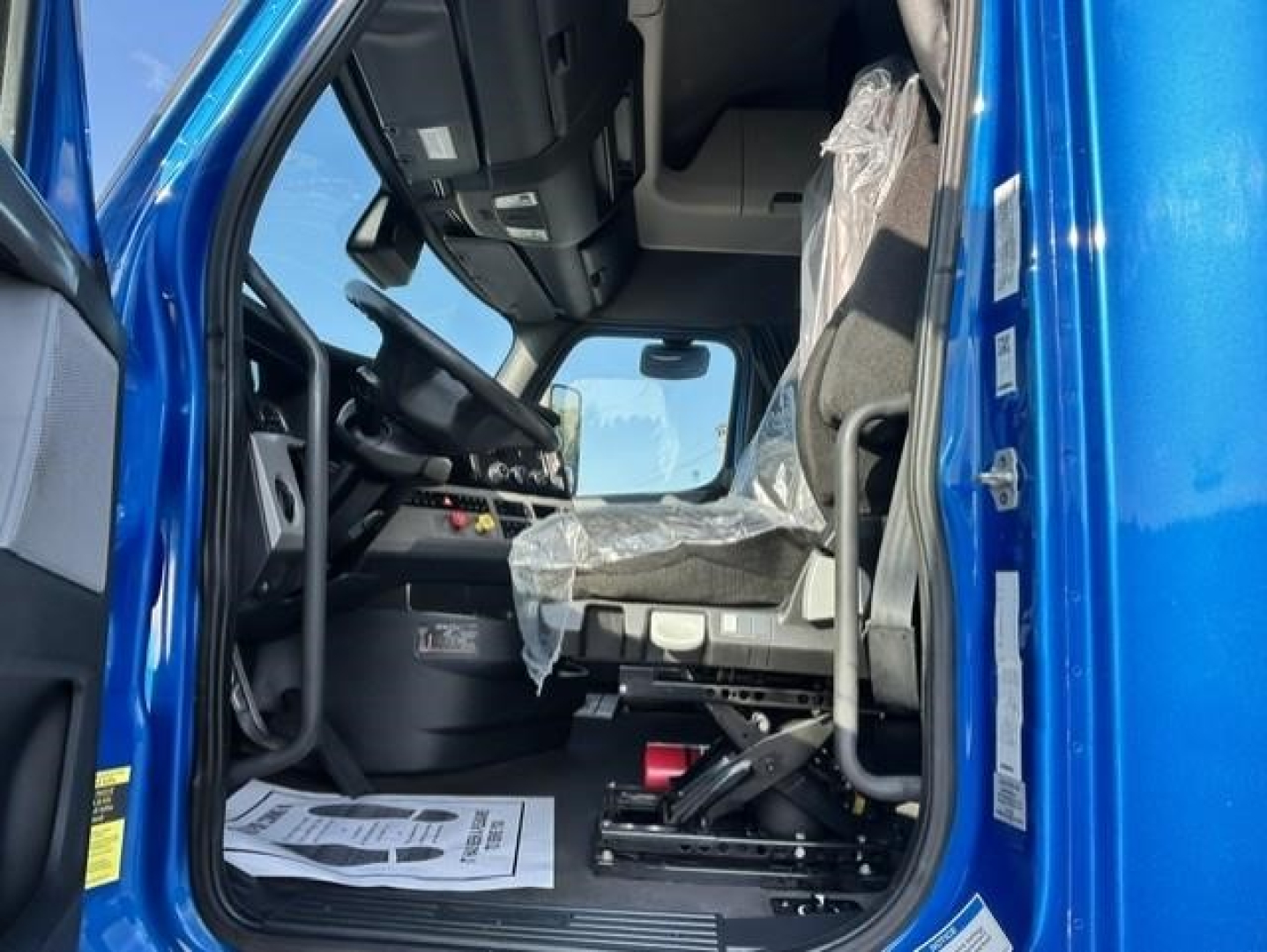 Truck cab interior of a blue 2022 Freightliner Cascadia 126, driver seat with plastic cover, steering wheel, dashboard, and entry door visible.