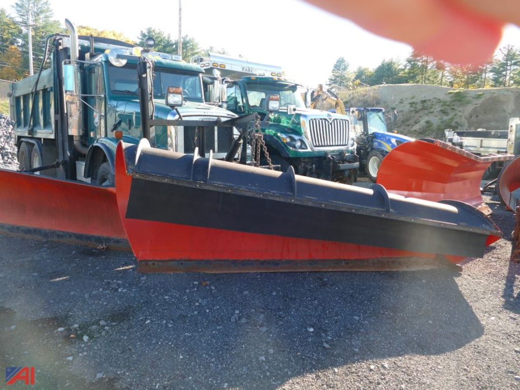 Freightliner truck chassis with a large red snow plow and wing attachment in a lot; other trucks visible in background.