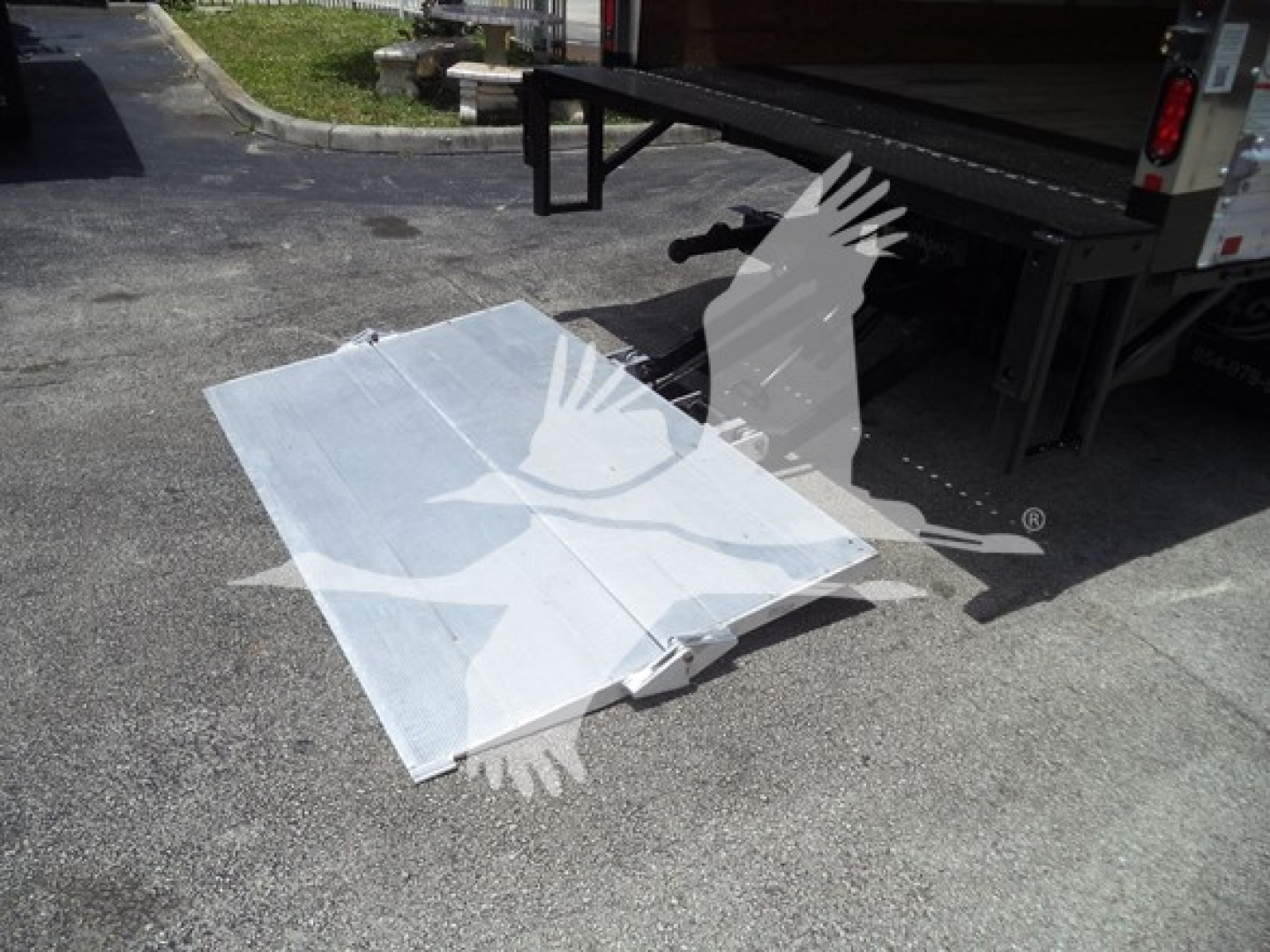 Isuzu NPR truck bed ramp folded on pavement adjacent to a flatbed trailer.