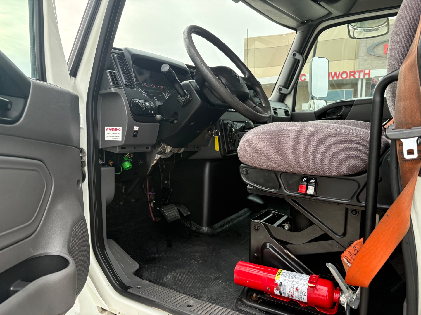 Truck cab interior of a 2020 International MV607 showing steering wheel, dashboard, and driver seat with orange seatbelt; partial view of door and pedals.