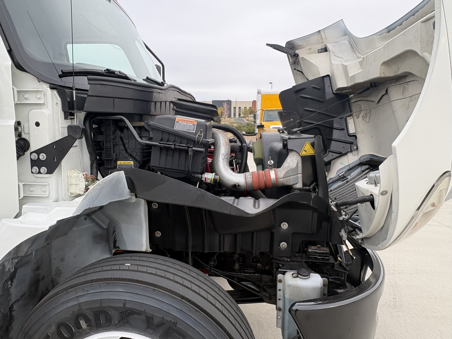 Truck engine bay of a 2020 International MV607 with hood open, showing cold storage refrigeration unit and engine components. Visible: radiator, hoses, and front wheel area.