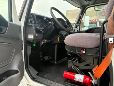 Truck cab interior of a 2020 International MV607 showing steering wheel, dashboard, and driver seat with orange seatbelt; partial view of door and pedals.