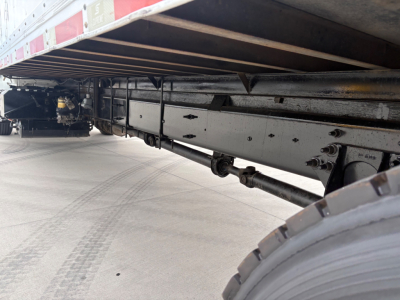 Underbody of a refrigerated truck trailer showing frame rails, suspension, and wheel, with drivetrain components visible toward the front of the trailer.