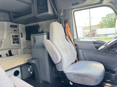 Truck cab interior showing driver seat and sleeper area with control panel, storage cabinet, and wood bed; light gray upholstery.