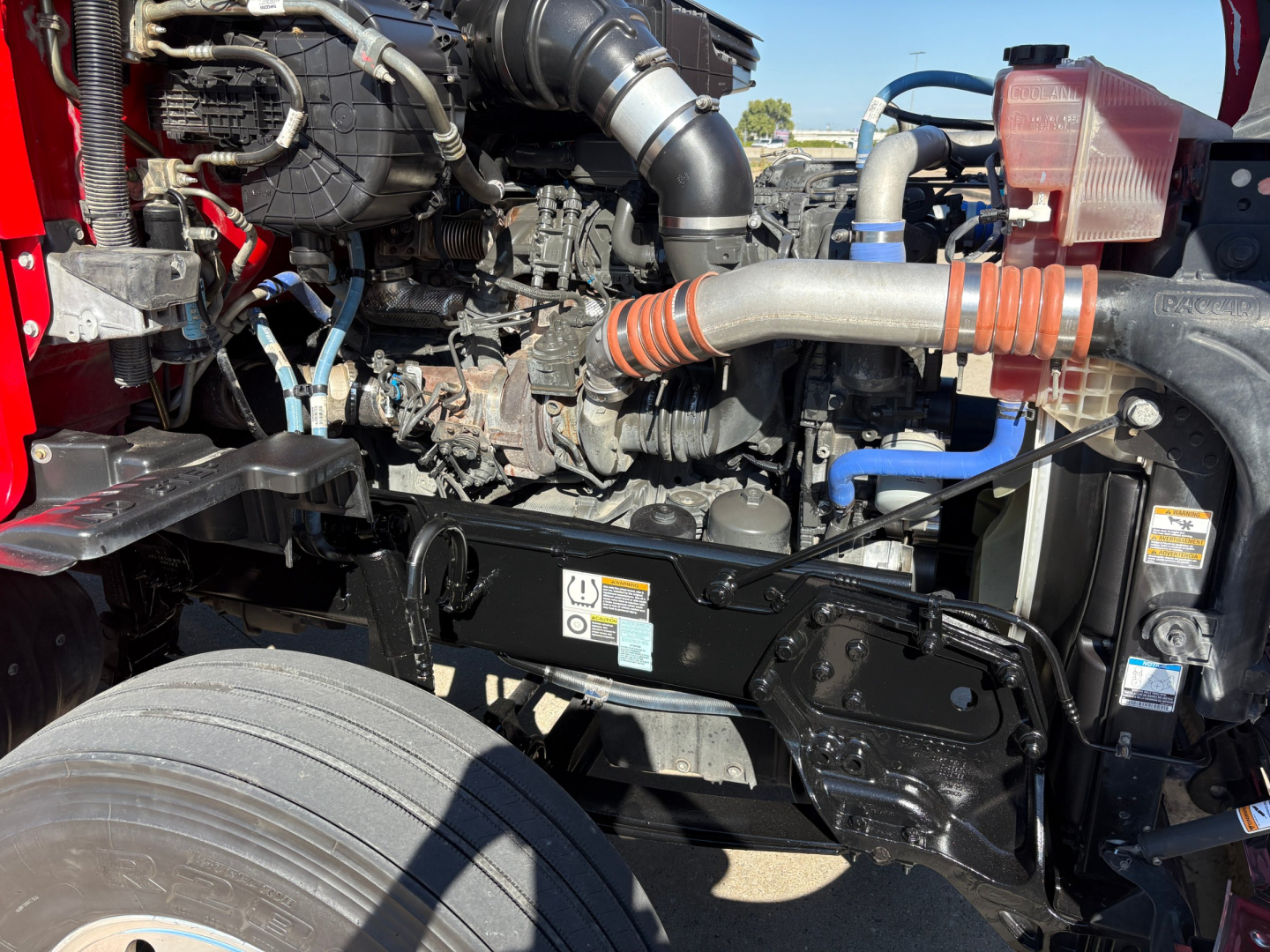 Kenworth T680 engine bay showing large V-engine with hoses, intercooler piping, radiator, and front suspension components.
