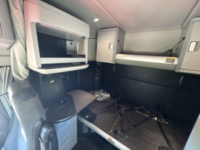 Kenworth T680 truck cab interior showing sleeper area with overhead cabinets, bunk bed, and a desk area; small items on the floor and a windowed shelf visible.