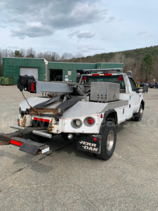 Tow truck rear view showing flatbed with metal ramps and equipment mounted on bed, white cab, black mud flaps, and dual rear wheels.
