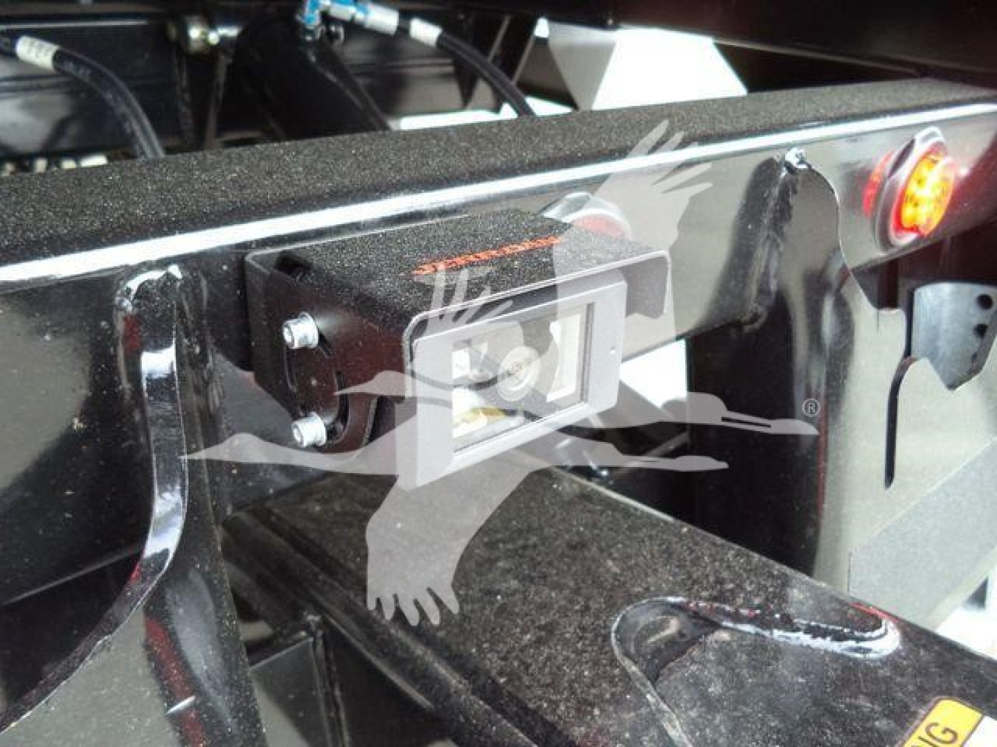 Tractor-trailer hitch area with a rectangular camera/monitor device mounted on a black metal frame, visible bolts, wiring, and a small orange label on top.
