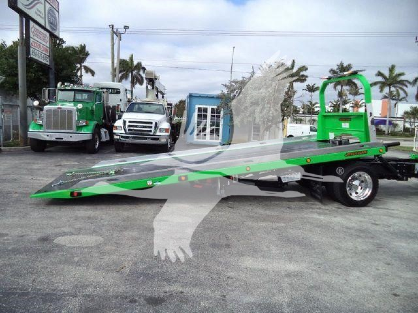 Light green Peterbilt rollback tow truck with a lowered flatbed and a green rollover wheel lift, parked in a lot with other trucks in background.