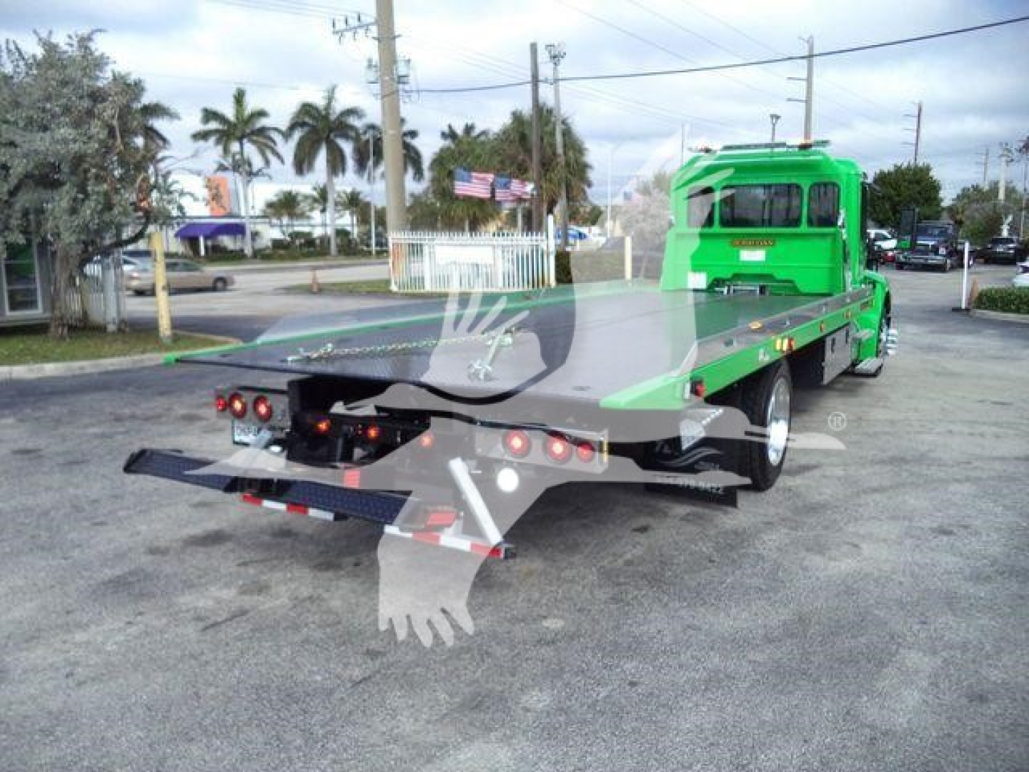 Green Peterbilt 536 rollback truck with flatbed, rear view showing taillights and ramps; license-free area with palm trees in background.