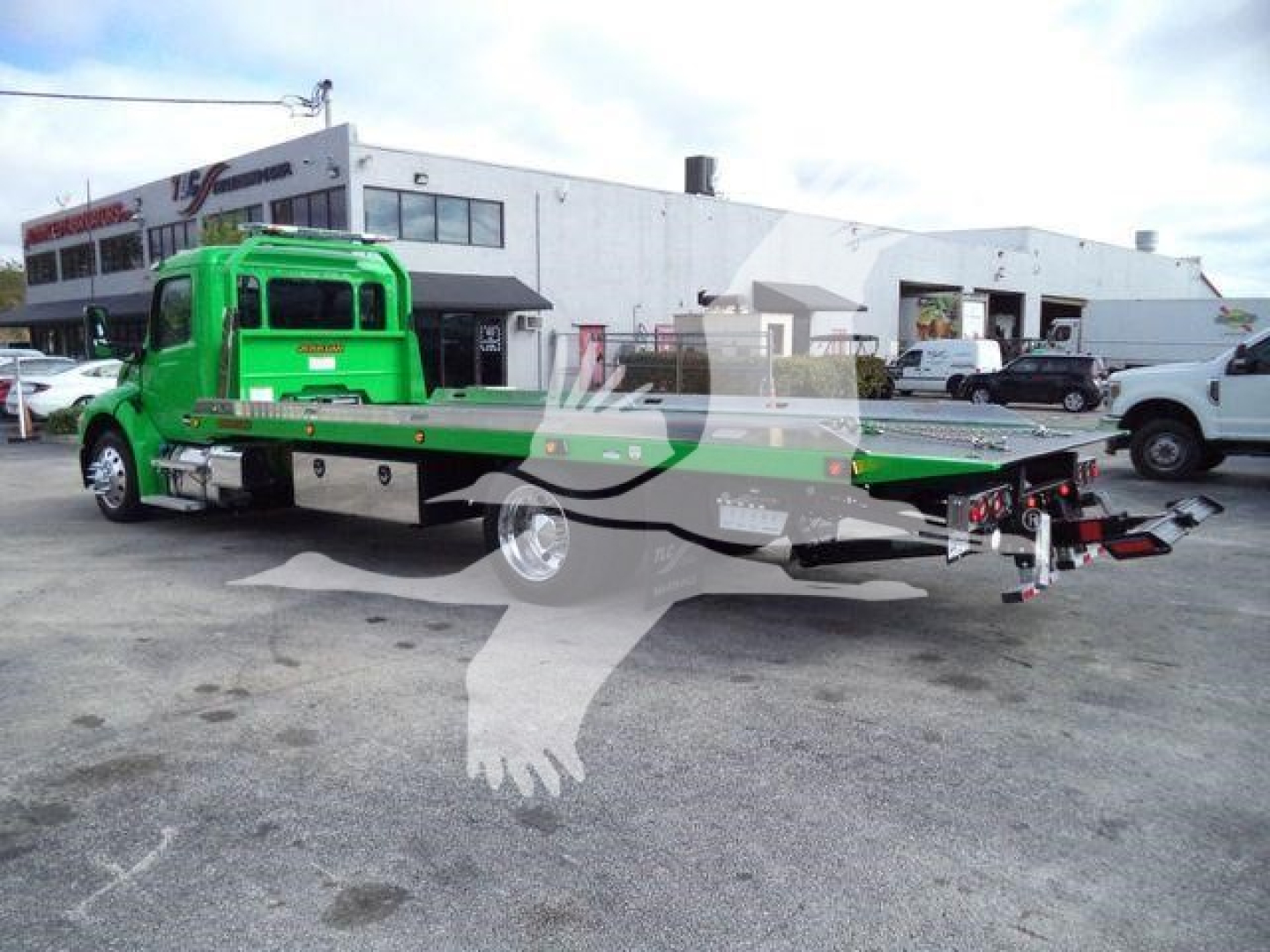 Green Peterbilt rollback tow truck shown in side view with flatbed extended, front cab visible, dealership in background.