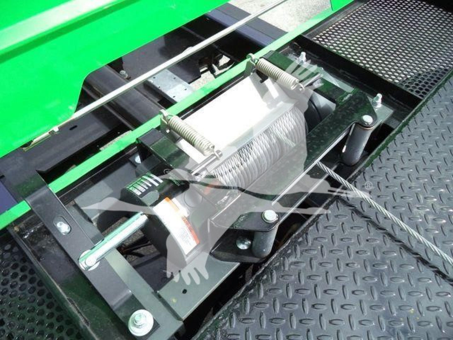 Equipment winch or hoist assembly mounted on a green Peterbilt 536 rollout truck, showing a metal winch drum with cable, bolts, and mounting brackets on a diamond plate surface.