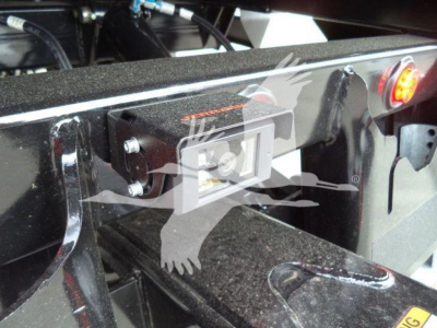 Tractor-trailer hitch area with a rectangular camera/monitor device mounted on a black metal frame, visible bolts, wiring, and a small orange label on top.