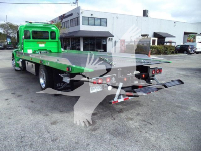 Green Peterbilt rollback truck with flatbed in a dealership lot. Visible text includes Peterbilt logo on cab, wheel chocks, and rear hydraulic ramps.