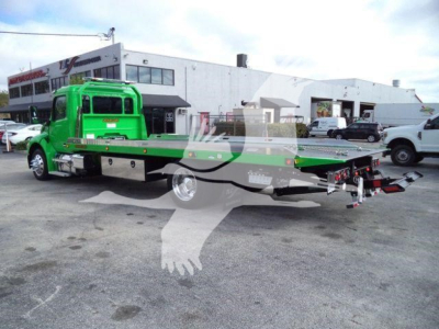 Green Peterbilt rollback tow truck shown in side view with flatbed extended, front cab visible, dealership in background.
