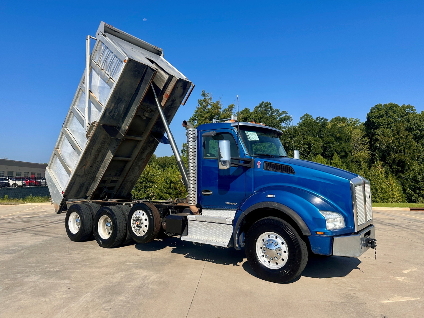 Kenworth T880 dump truck in blue cab with raised silver dump bed, three-axle configuration, side view.