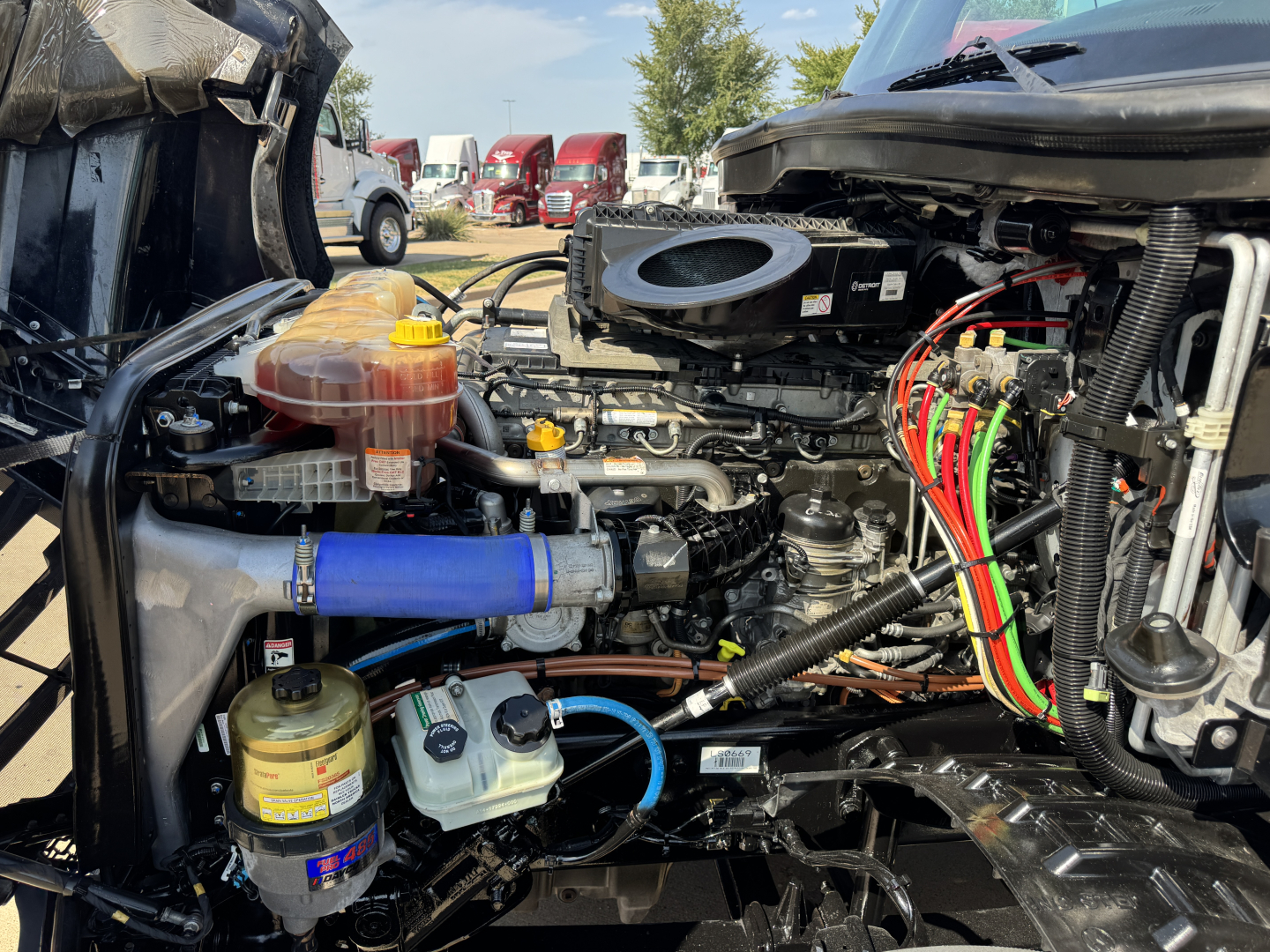 Truck engine bay with visible fluid reservoirs, hoses, and wiring harness; blue silicone hose, coolant reservoir, power steering fluid container, and air intake components visible.