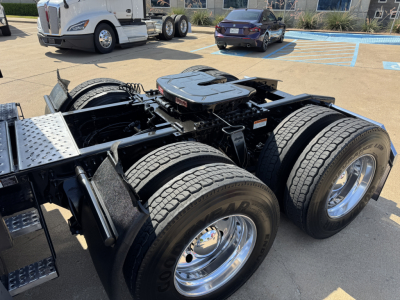 Truck chassis with dual rear axles and fifth wheel mounting in foreground; tandem axle setup, fuel tanks, and cab in distance.