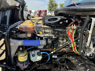 Truck engine bay with visible fluid reservoirs, hoses, and wiring harness; blue silicone hose, coolant reservoir, power steering fluid container, and air intake components visible.