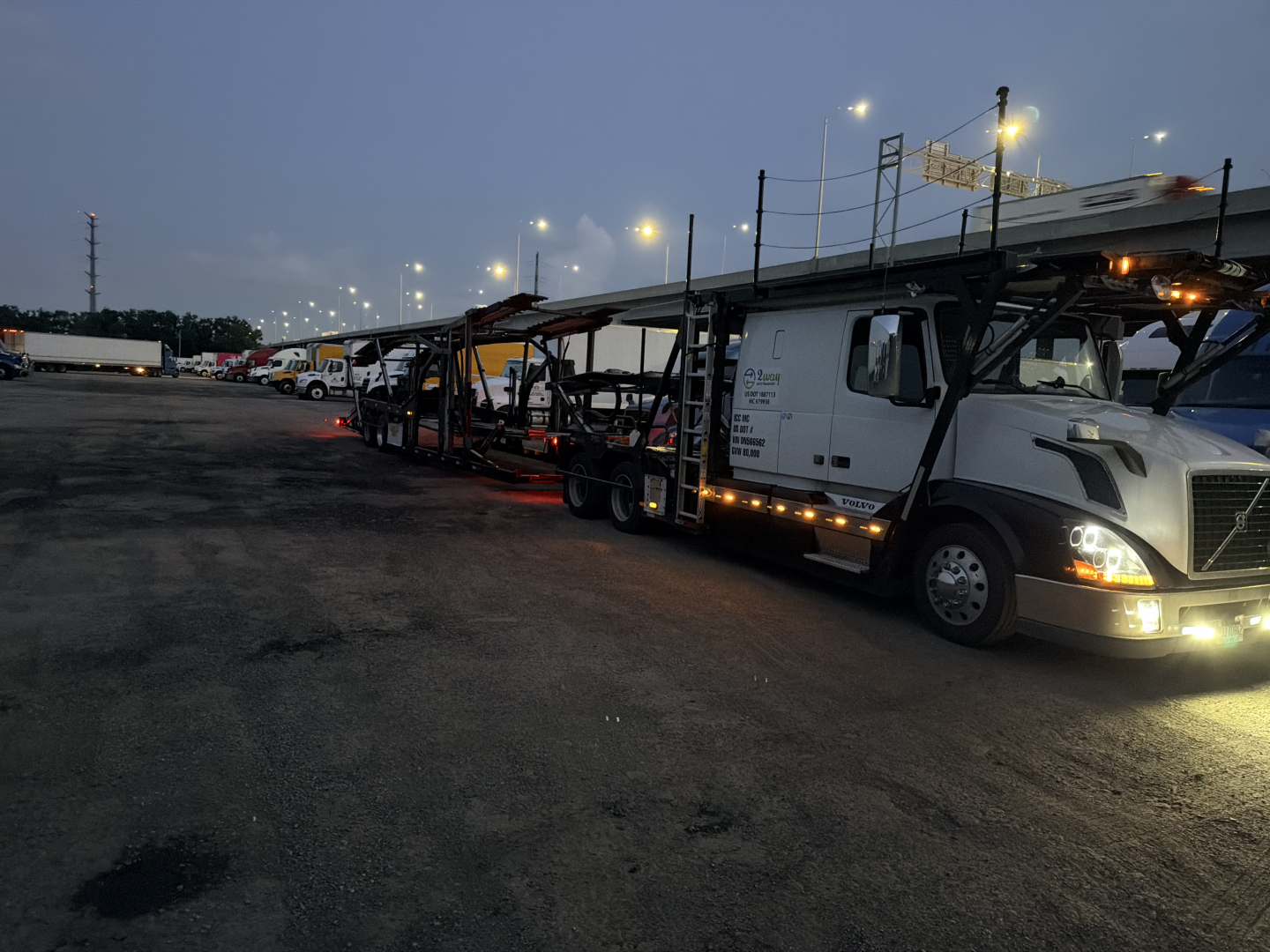Semi truck in a lot showing a Volvo VNL cab and chassis loaded on a car carrier trailer with lights on at dusk.